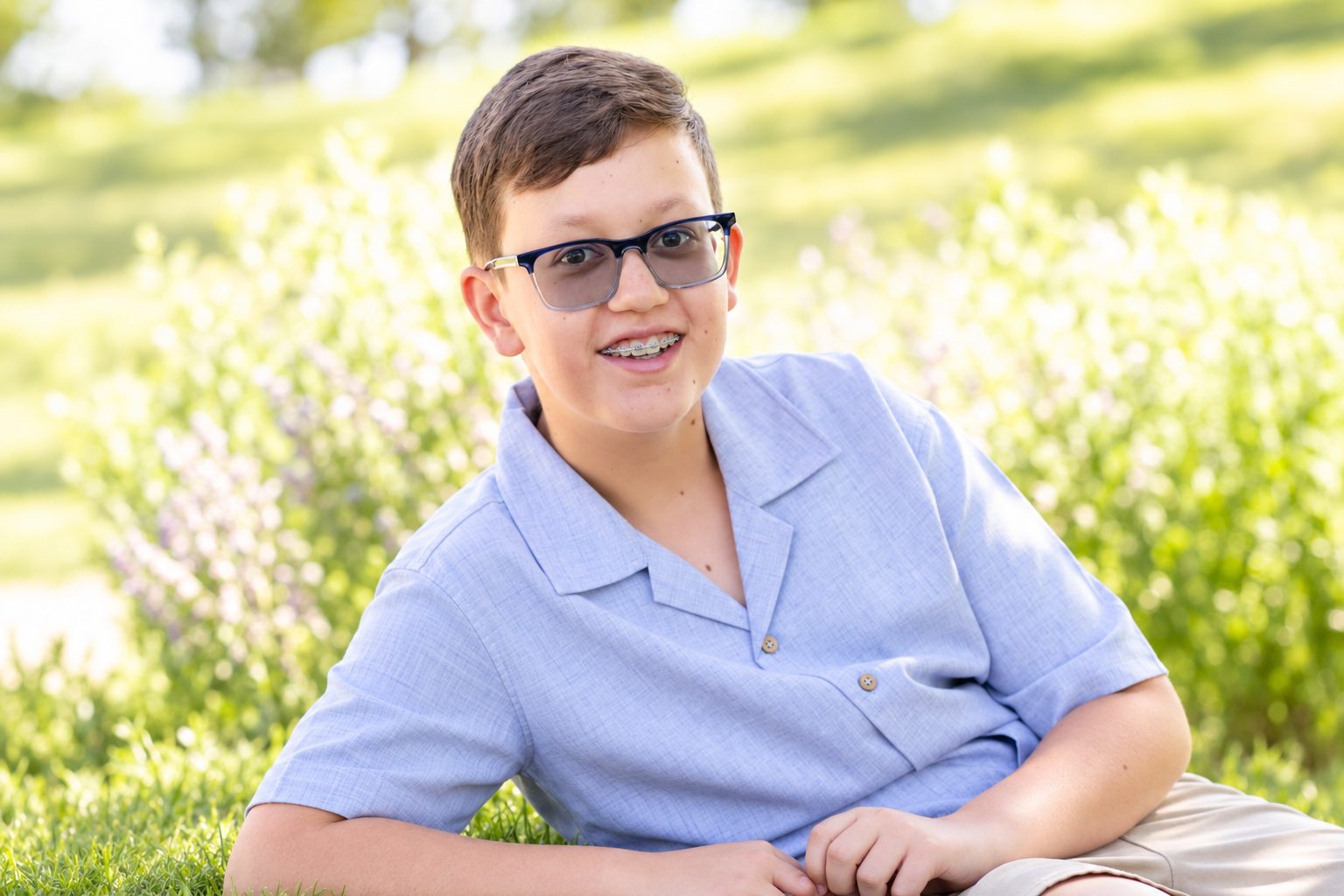 A smiling teenager with braces and glasses, sitting on the grass outdoors.